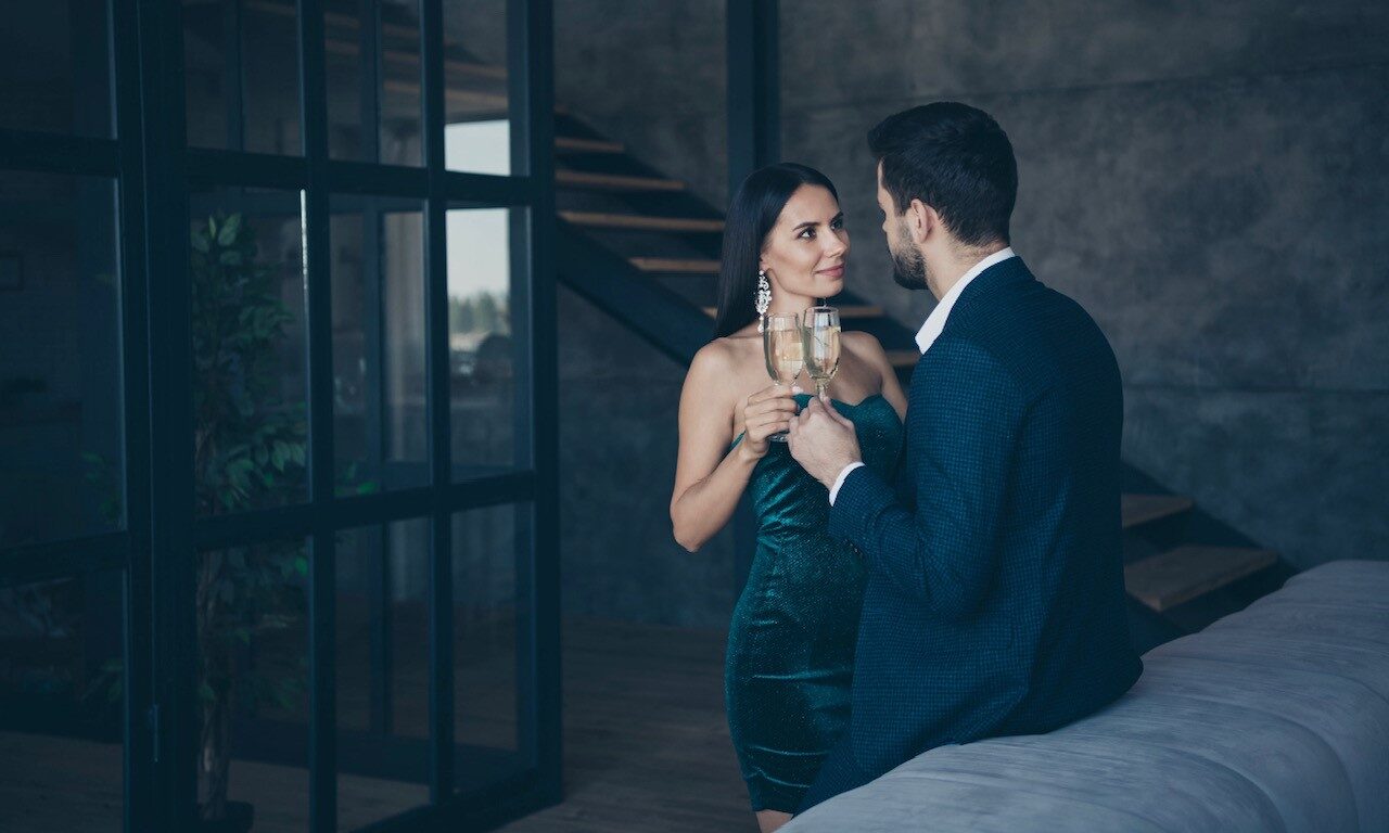 Successful couple in classy attire standing inside a multi million dollar house drinking wine.